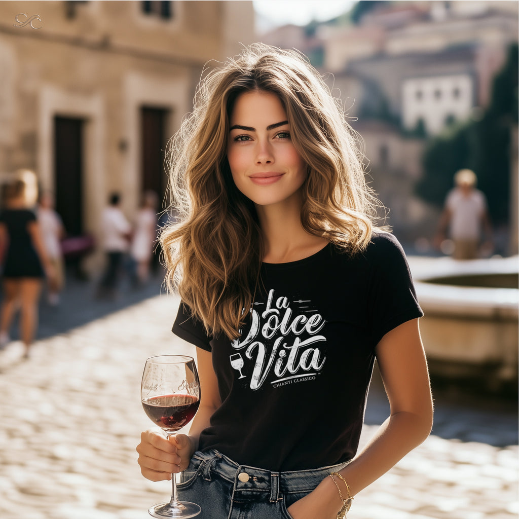 Model wearing black ‘La Dolce Vita’ Chianti Classico graphic tee, holding a glass of red wine while standing in a sunlit cobblestone street with historic buildings in the background.
