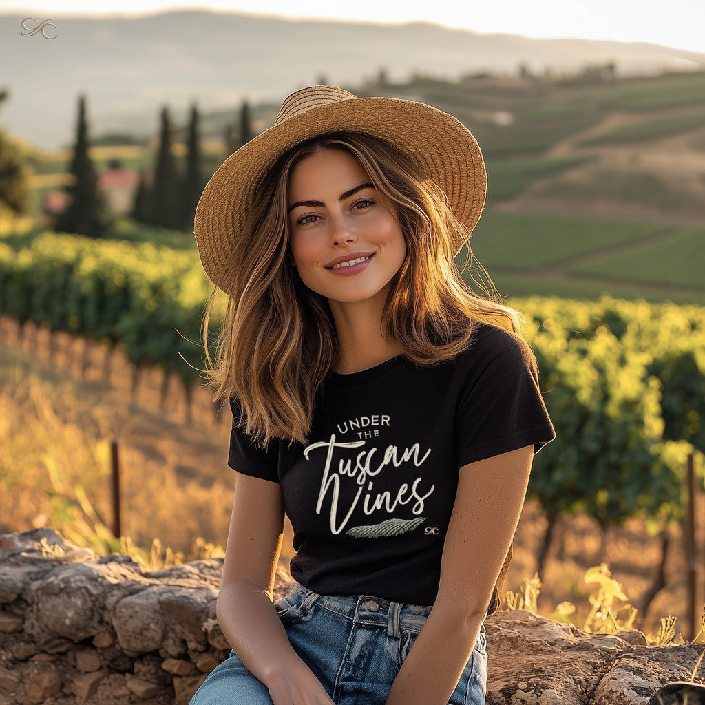 Woman in a straw hat sitting by a vineyard wall at sunset, wearing a black tee with the phrase "Under the Tuscan Vines" in script across the chest.
